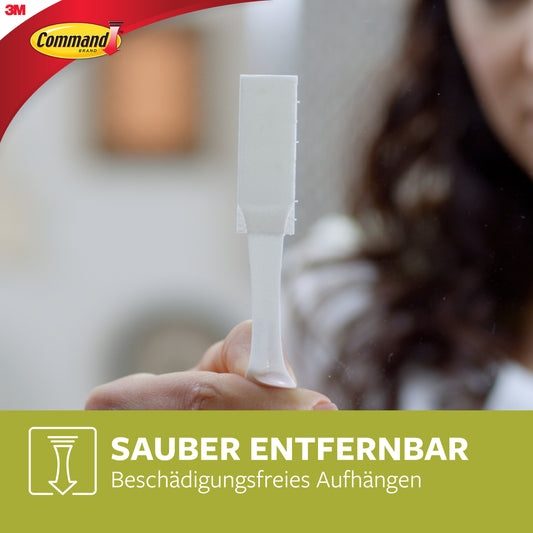 Eine Person hält einen weißen Klebestreifen mit dem Logo der 3M Deutschland GmbH. Der Text lautet: "Sauber entfernbar - Bilder aufhängen ohne Nägel mit mittelgroßen Command™ Bilder-Montage Strips M 17201-4PKFGN.