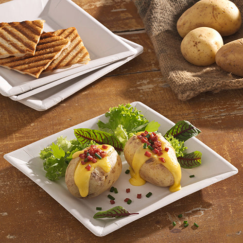 Zwei gebackene Kartoffeln mit gelber Käsesauce, Speckstückchen und Schnittlauch werden auf einem PAPSTAR Papp-Teller eckig 22,5 cm im "pure" Design von PAPSTAR GmbH serviert. Blattgemüse begleitet das Gericht, im Hintergrund stehen gegrilltes Brot und ganze Kartoffeln.