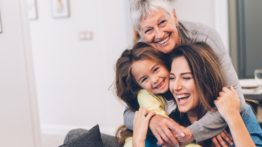 Das Bild zeigt drei Generationen von Frauen – eine ältere Dame, eine Mutter und ihre Tochter. Alle drei sind in einem hellen, freundlichen Raum zu sehen und umarmen sich glücklich. Die ältere Dame lacht und ist hinter der Mutter und Tochter, die ebenfalls lächeln und sich in der Mitte des Bildes befinden. Die Atmosphäre vermittelt Liebe, Freude und familiäre Nähe. Es scheint ein besonderer Moment des Zusammenseins und der Zuneigung zu sein.
