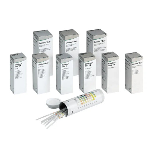 Hinter einem offenen zylindrischen Behälter mit Deckel verbergen sich neun Schachteln mit der Aufschrift „Combur®“ der Servoprax GmbH, die jeweils 100 Roche Urinteststreifen enthalten. An der Seite des Behälters ist eine mehrfarbige Tabelle angebracht, die sich perfekt für die Durchführung eines Schnelltests eignet.