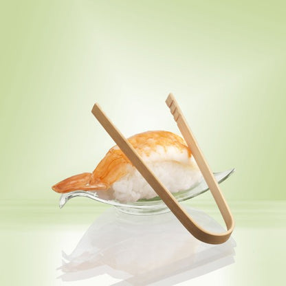 A shrimp nigiri sushi is served on a glass dish, complemented by PAPSTAR bamboo finger food tongs (10 cm, 50 pieces) from PAPSTAR GmbH. A gentle light green highlights the eco-friendly bamboo presentation.