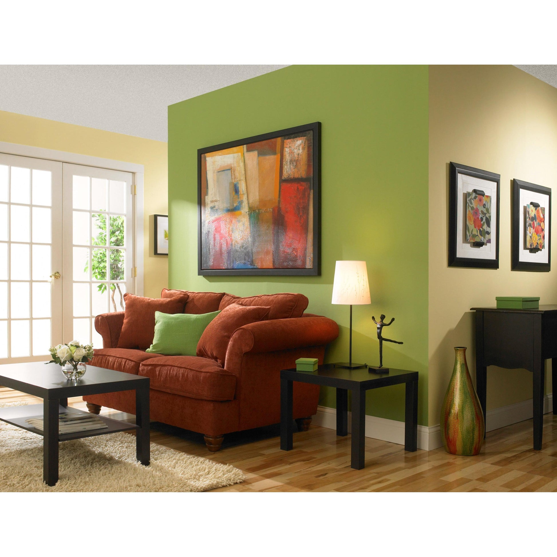 Ein Wohnzimmer mit gelben und grünen Wänden, die mit ScotchBlue™ Malerabdeckband "Multi-Surface" von 3M Deutschland GmbH gestrichen sind, einem roten Sofa mit grünen Kissen, einem schwarzen Couchtisch, einem Beistelltisch mit Lampe, abstrakter Kunst, Holzboden und Glastüren.