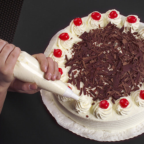 With the Starpak 10 piping bag 1.8 l with 3 piping nozzles, a round cake is decorated with piped white cream, chocolate shavings in the middle, and cherries on top, placed on a white cake doily on a dark background.