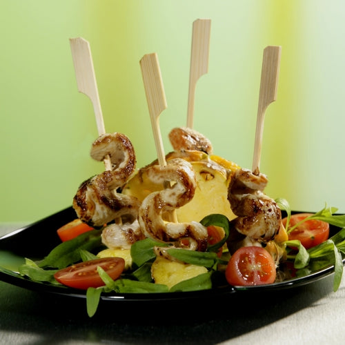 A black plate with salad, halved cherry tomatoes, pineapple, and PAPSTAR GmbH finger food skewers "pure" 10 cm (40 pieces) - grilled chicken skewers arranged vertically. Ideal for golf events and catering. The green background is blurred.