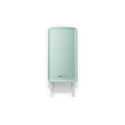 The CWS PureLine front panel for the toilet paper dispenser from CWS, depicted in mint green with a visible leaf hanging down from below, is mounted on the wall and photographed against a white background to achieve a hygienic look.