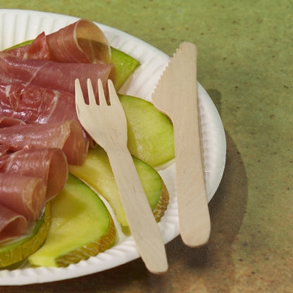 A paper plate with sliced green apples and rolled cured meat rests alongside a PAPSTAR wooden knife "pure" 16.5 cm from PAPSTAR GmbH and a wooden fork on a green surface.