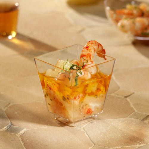 Eine Starpak 50 Fingerfood-Schale (PS glasklar) von Starpak, gefüllt mit Garnelen und Meeresfrüchtecocktail und garniert mit Kräutern, steht auf einem beigefarbenen, strukturierten Untergrund. Weitere Tassen sind im Hintergrund verschwommen – perfekt für stilvolle Events.