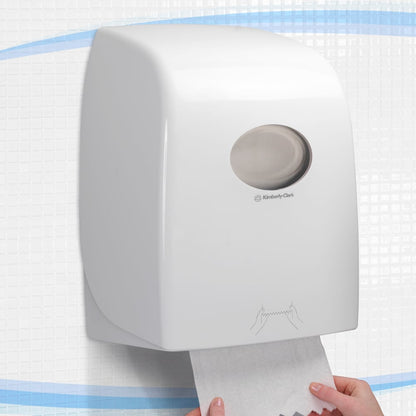 Der Aquarius™ Rollenhandtuchspender - Weiß von Kimberly-Clark GmbH wird an der Wand auf Fliesen montiert, wobei eine Hand ein Handtuch von unten zieht. Er ist ideal für öffentliche Bereiche und verfügt über ein rundes Fenster oben und eine klare Gebrauchsanweisung darunter.