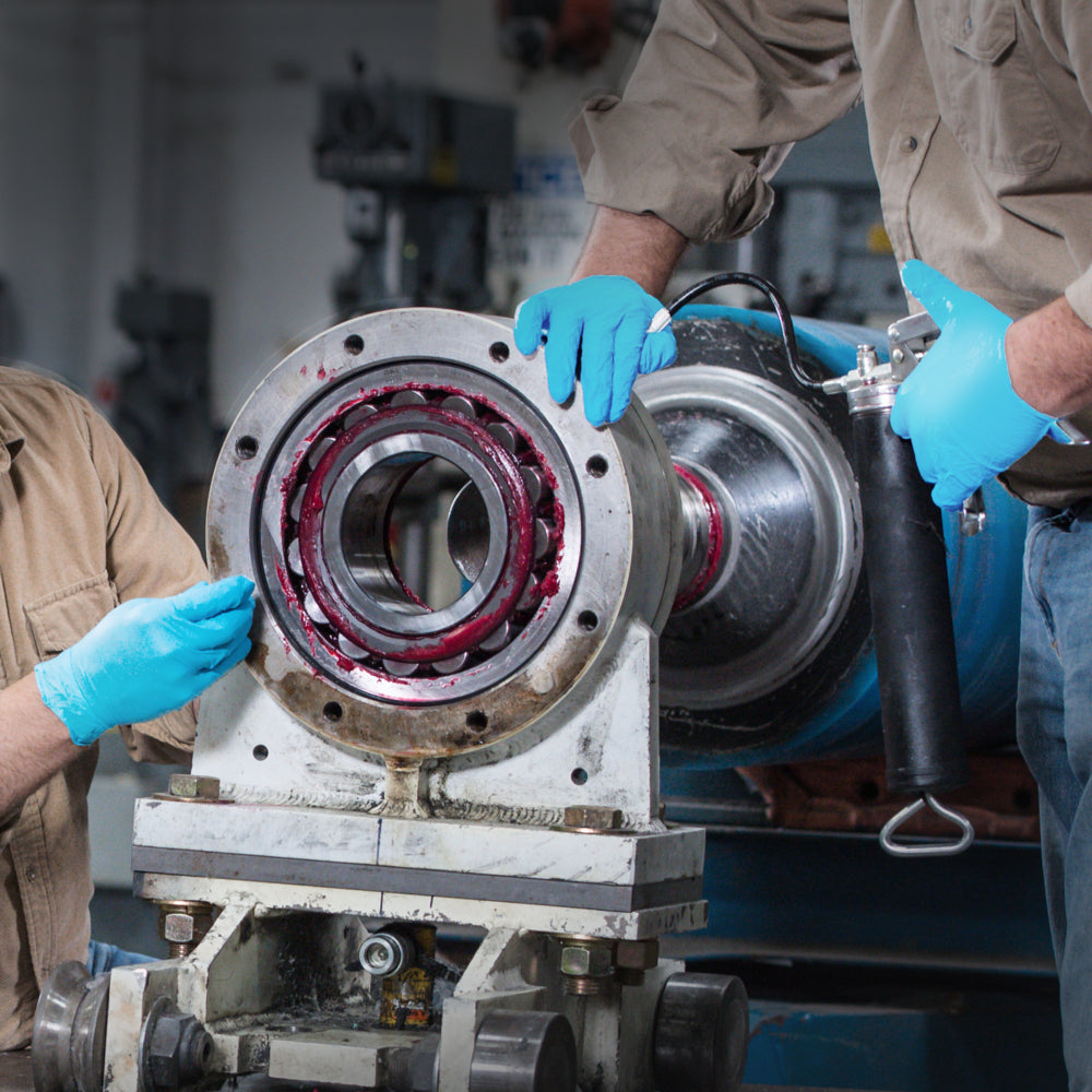 Zwei Personen in hellbraunen Hemden tragen Kimberly-Clark GmbH KleenGuard® G10 Comfort Plus Blaue Nitril-Handschuhe (24 cm, beidhändig tragbar), während sie mit einer Fettpresse Schmiermittel auf ein Lager einer Industrieanlage in einer Werkstatt auftragen.