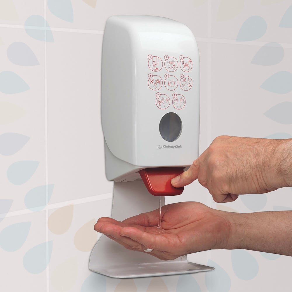 Eine Person drückt einen roten Hebel an einem an der Wand montierten Kleenex® Extra Gel Händedesinfizieren-Spender (Kimberly-Clark GmbH) und gibt klares Händedesinfektionsmittel auf ihre Hand ab. Auf der Vorderseite ist eine illustrierte Anleitung zu sehen, dahinter befinden sich gemusterte Kacheln.