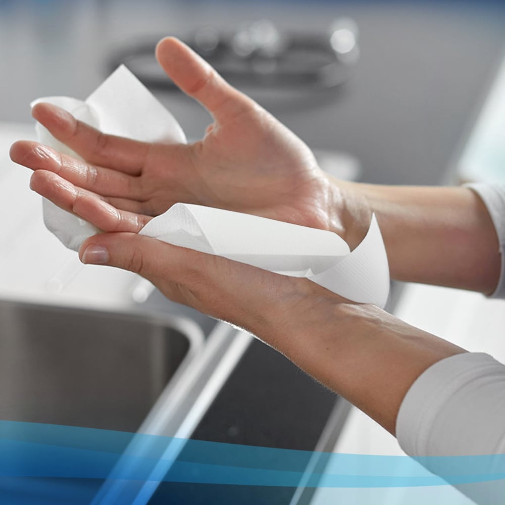 Jemand trocknet sich die Hände mit Scott® Control™ Handtüchern - Interfold / Weiß / Klein von Kimberly-Clark GmbH, mit einer Küchentheke und einem Wasserhahn im Hintergrund. Die Packung enthält 15 x 304 Blatt pro Karton.