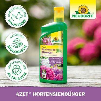 Eine grüne Flasche Neudorff Shop Azet HortensienDünger flüssig mit Blumenbildern auf dem Etikett, abgebildet mit Symbolen für pet-safe, organischer Dünger und Biokunststoff. Der Hintergrund zeigt verschwommene Blumen.