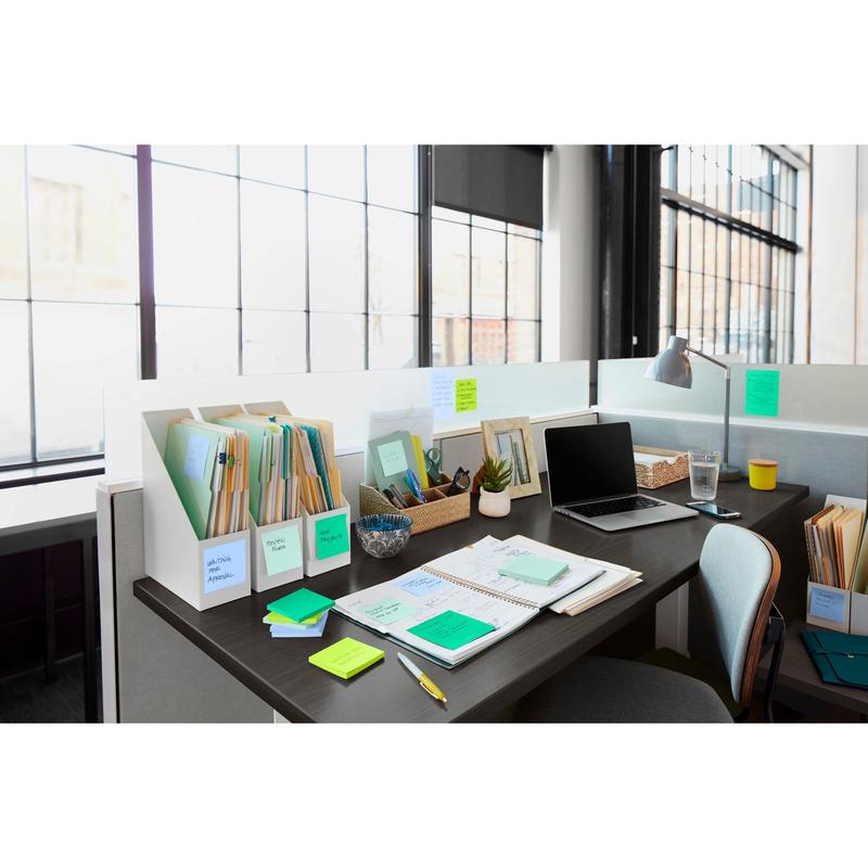 Ein heller Bürotisch mit Ordnern, Notizbüchern, Schreibwaren, einem 3M Deutschland GmbH Post-it® Super Sticky Notes Block (76x127mm, 90 Blatt, 100% PEFC), einer kleinen Pflanze, einer Lampe, einem Glas Wasser und einem offenen Laptop an einem großen Fenster.