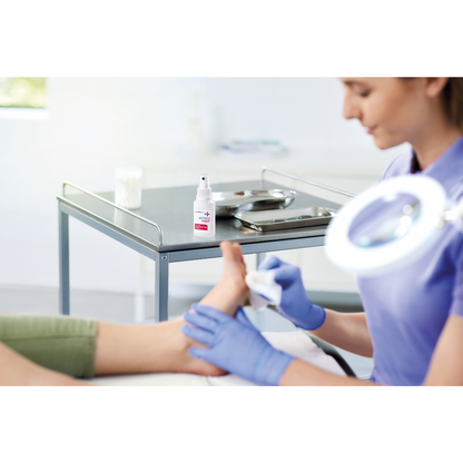 A person with blue gloves is cleaning another person's foot in a clinic. The focus is on infection protection. On the table are a 50 ml bottle of Schülke octenisept® wound disinfection spray from Schülke & Mayr GmbH and a metal tray with tools.