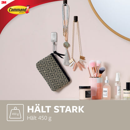 Ein kleiner Command™ Metallhaken in gebürstetem Nickel (3M Deutschland GmbH) hält ein gemustertes Täschchen und Schmuck an einer rosa Wand. Darunter auf dem Tisch: Make-up-Pinsel, Kerze und Kosmetik. Text: "HÄLT STARK Hält 450 g.