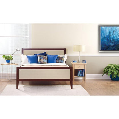 Ein ordentlich gemachtes Bett mit weißen und blauen Kissen steht vor einer hellen Wand, die mit ScotchBlue™ Malerabdeckband "Multi-Surface" der 3M Deutschland GmbH beklebt ist. Auf einem Nachttisch stehen eine Lampe, eine Uhr, Bücher und eine Topfpflanze neben dem hellen Fenster.