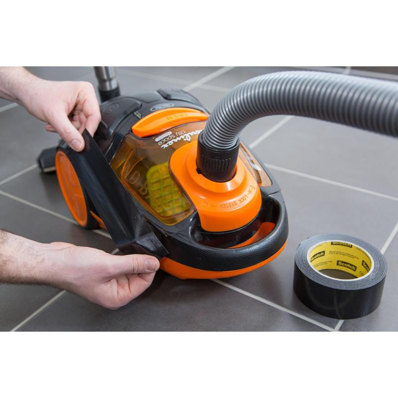 Eine Person klebt Scotch® Universal Klebeband 2904 von 3M Deutschland GmbH auf die beschädigte Seite eines orange-schwarzen Staubsaugers auf einem grauen Fliesenboden, während eine Rolle des Klebebands in der Nähe liegt.