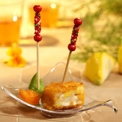 On a transparent, leaf-shaped plate lies a cube of breaded, deep-fried food with a PAPSTAR 250 finger food picks from the PAPSTAR GmbH, along with a green slice and an orange slice. Against the slightly blurred background, you can see a slice of lemon and some greens.