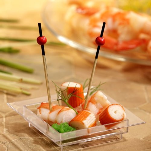 On a transparent, square plate are imitation crab, shrimp, a green garnish, and two PAPSTAR 250 finger food picks from the PAPSTAR GmbH with red beads. In the background, blurry shrimp can be seen on a plate.