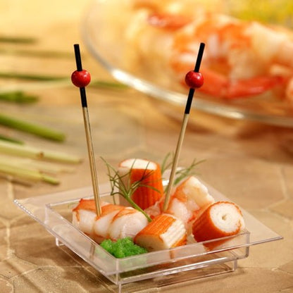 On a transparent, square plate are imitation crab, shrimp, a green garnish, and two PAPSTAR 250 finger food picks from the PAPSTAR GmbH with red beads. In the background, blurry shrimp can be seen on a plate.