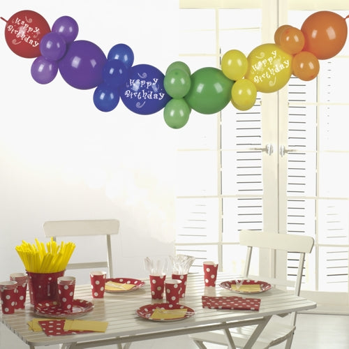 A white table with red polka-dotted partyware, yellow utensils, and cups is crowned by the PAPSTAR balloon garland 2 m "Happy Birthday" from PAPSTAR GmbH - a festive birthday party decoration that shines in the sunlight through white shutters.