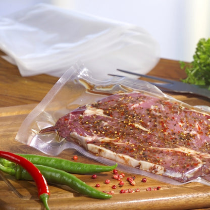 Raw, seasoned meat is vacuum-sealed in a transparent PAPSTAR 100 edge-sealed bag (PA/PE) from PAPSTAR GmbH on a wooden cutting board, surrounded by chili peppers, peppercorns, cutlery, and leafy vegetables.