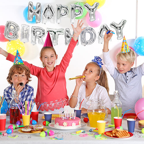 Four children with party hats celebrate a birthday at a decorated table with cake and party items. Behind them, the "Happy Birthday" foil balloon set from PAPSTAR GmbH in silver and colorful balloons decorate the wall.