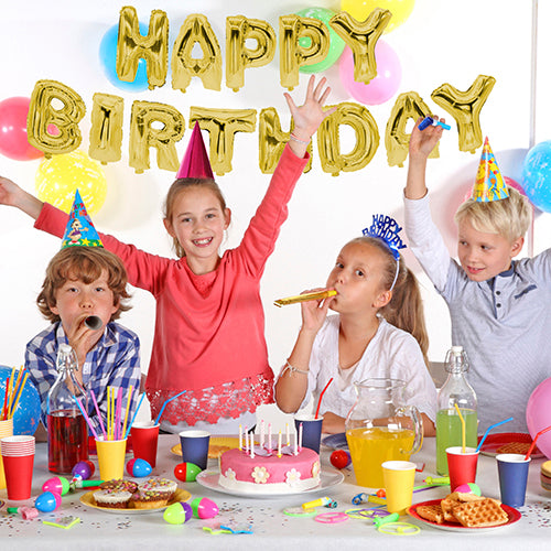 Four children with party hats sit at a festively set table with cake and drinks. On the wall, the "Happy Birthday" foil balloon set from PAPSTAR GmbH adds a golden shine to the birthday celebration.
