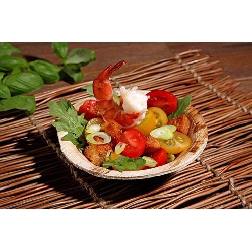Ein kleiner Salat mit Garnelen, Kirschtomaten, Rucola, Frühlingszwiebeln und Croutons wird in einer PAPSTAR 6 Schalen Palmblatt „pure“ rund 200 ml Schüssel (Ø 13 cm · 3 cm) der PAPSTAR GmbH auf einem gewebten Tischset mit Basilikumblättern im Hintergrund serviert.