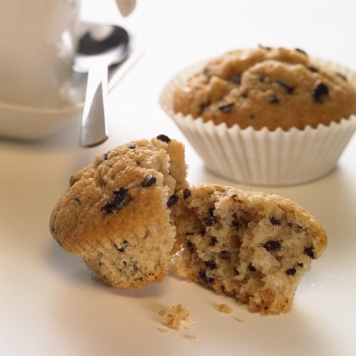 Ein Schokoladenmuffin liegt halbiert auf einer weißen Fläche, daneben befinden sich Krümel. Im Hintergrund liegt ein weiterer ganzer Muffin in einem PAPSTAR 1000 Backförmchen rund der PAPSTAR GmbH, daneben ein Teil einer Kaffeetasse und ein Löffel.