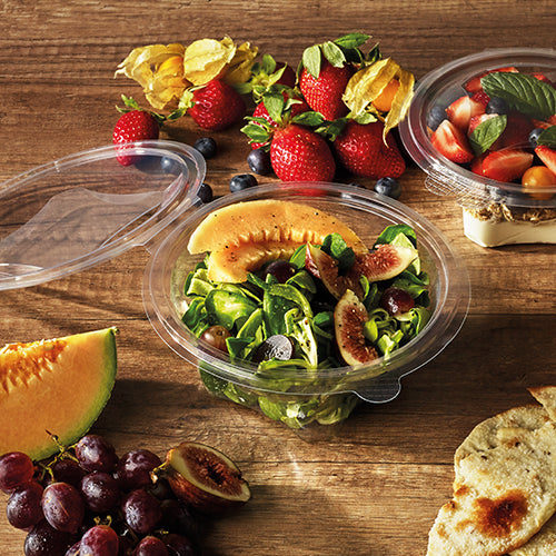 On a wooden table stands a round Starpak 50 delicatessen and salad bowl with hinged lid (R-PET crystal clear) with a salad of melon, figs, and leafy greens. Next to it are strawberries, grapes, melon slices, cherry tomatoes, flatbread, and another bowl with tomatoes and basil.