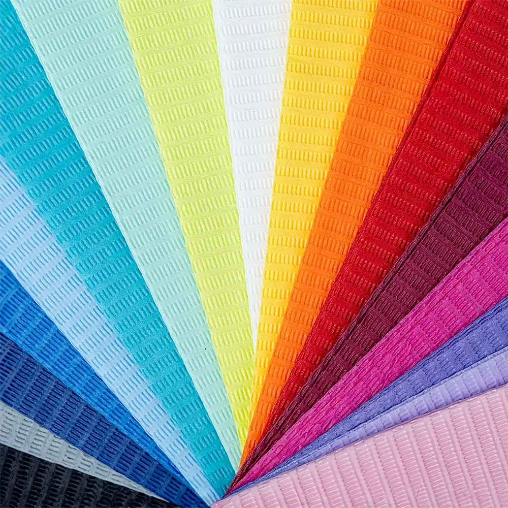 A series of AMPri Dental disposable patient napkins from AMPri Handelsgesellschaft mbH, arranged in a fan shape, shows bright colors like light blue, teal, green, yellow, red, orange, pink, blue, purple, black, and white. Each napkin features a grid-like embossing pattern reminiscent of textured plastic foils used in dental treatments.