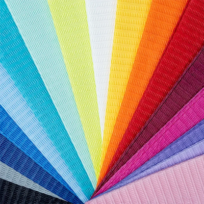 A series of AMPri Dental disposable patient napkins from AMPri Handelsgesellschaft mbH, arranged in a fan shape, shows bright colors like light blue, teal, green, yellow, red, orange, pink, blue, purple, black, and white. Each napkin features a grid-like embossing pattern reminiscent of textured plastic foils used in dental treatments.