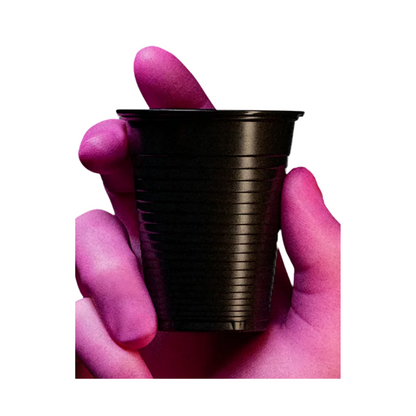 A person with purple-stained hands holds a small black AMPri Dental PP mouth rinse cup, manufactured by AMPri Handelsgesellschaft mbH, made of polypropylene between thumb and forefinger against a white background.