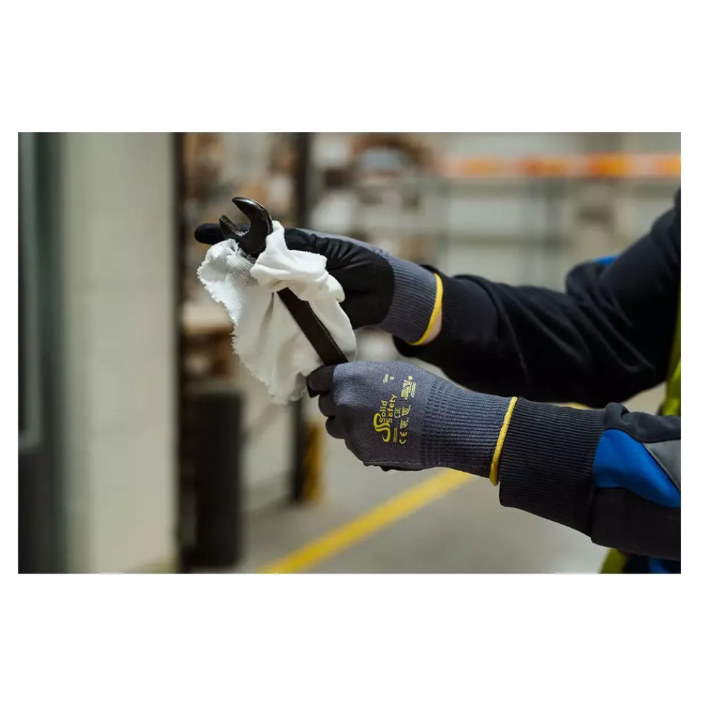 A person wearing AMPri SolidSafety Tough Air ultra-light assembly gloves from AMPri Handelsgesellschaft mbH carefully cleans a wrench indoors with a white cloth. The gray gloves, renowned for their abrasion resistance level of 4, stand out against the blurred industrial shelves and equipment in the background.