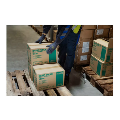 A person wearing AMPri SolidSafety Tough Micro Touch ultra-light assembly gloves is carefully stacking boxes labeled 'Basic-Touch Nitrile' on a wooden pallet in a warehouse. Several other boxes are visible, and the person seems to be organizing the inventory with precision and care.