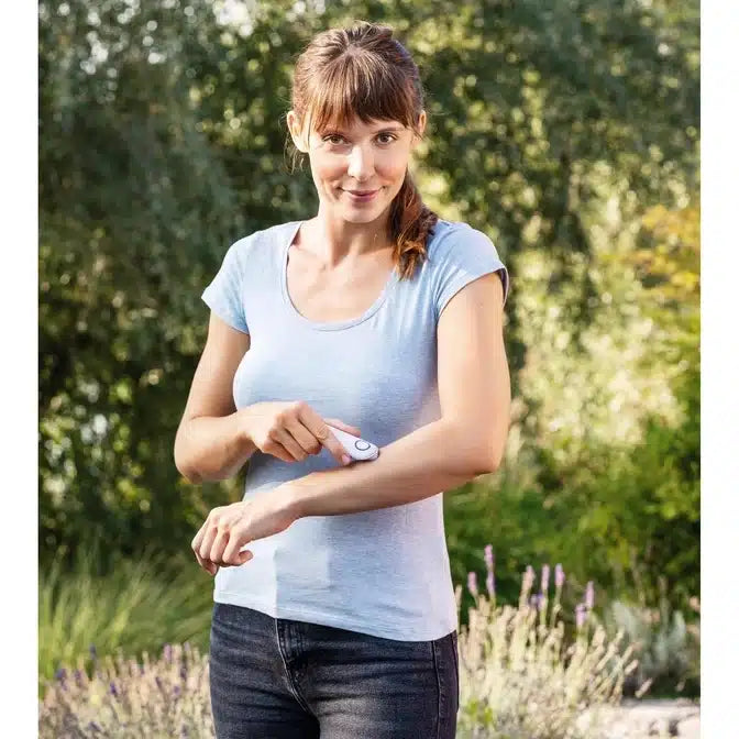 Eine Frau im hellblauen T-Shirt steht im Freien und checkt einen weißen Fitness-Tracker an ihrem Handgelenk. Im Hintergrund sind Laub und Sonnenlicht zu sehen, scheinbar unberührt von einem Insektenstichheiler BR 10 der Beurer GmbH.