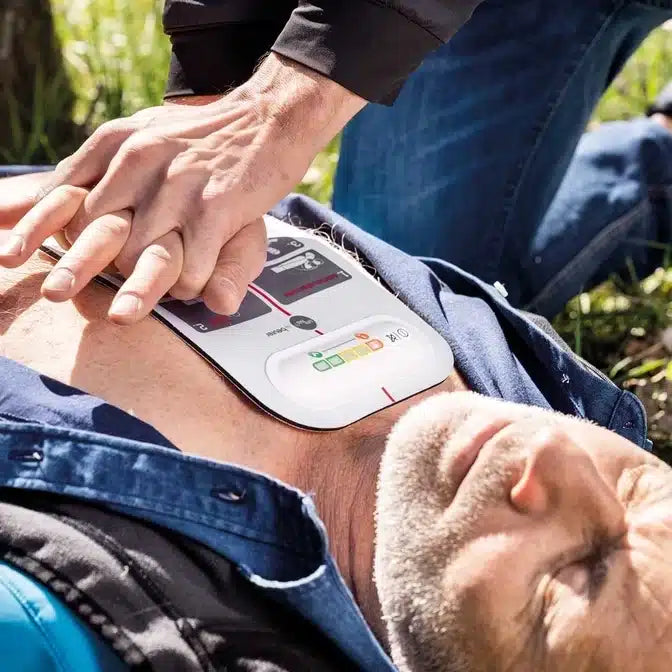 Ein erwachsener Mann liegt bewusstlos im Gras, auf seiner Brust liegt eine innotas Beurer LifePad® Reanimationshilfe, und die Hände einer Person drücken in einer Notfallsituation auf das Gerät.