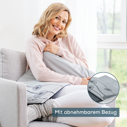 A woman sits on a light grey couch next to a window, holding the Beurer HK 125 XXL Heating Pad Cosy Grey from Beurer GmbH. She is smiling and wearing a pink hoodie. There is a close-up of the pad showing its removable cover and heating functions with 6 heat settings. The German text reads "With removable cover."