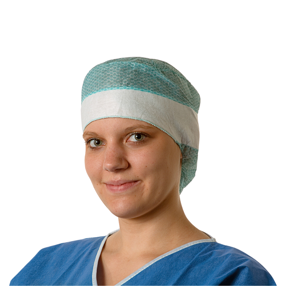A young doctor is wearing a blue surgical gown and a Hartmann Foliodress® Cap Comfort Form Plus disposable surgical cap made of breathable viscose fleece and is gently smiling at the camera. The background is plain white.
