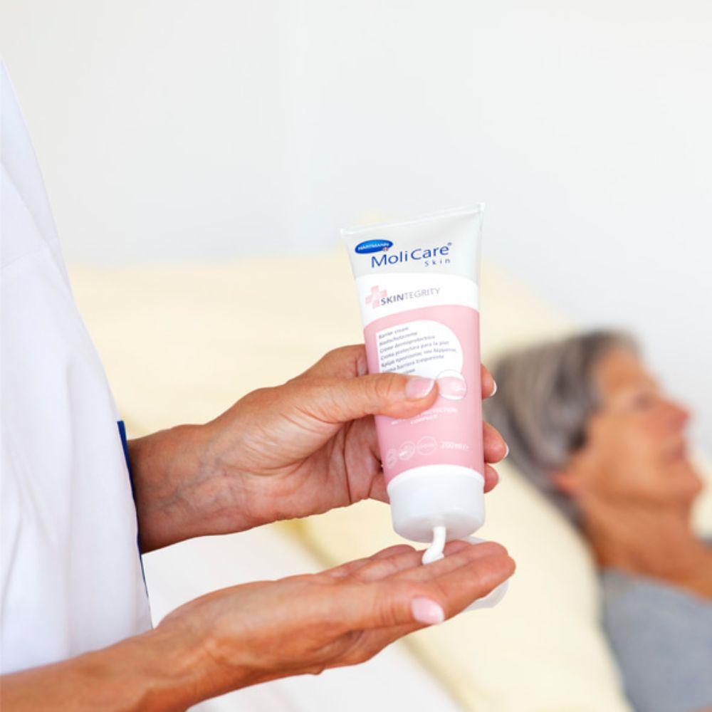 A person applies Hartmann MoliCare Skin Protective Cream (200 ml) by Paul Hartmann AG, enriched with Nutriskin Protection Complex, into their palm. In the blurred background, a small, gray-haired person lies on a bed, conveying the impression of personal care or a medical setting.