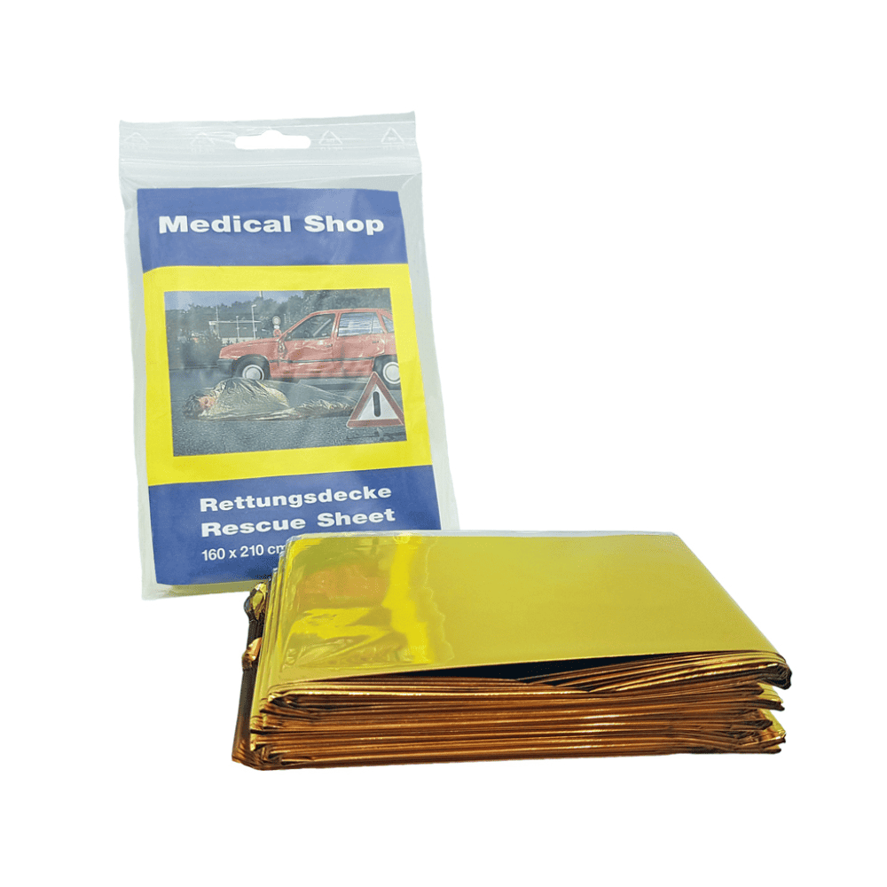 In front of it lies a folded, yellow-golden rescue blanket made of aluminum-coated polyester film. Behind it is a package labeled “Holthaus Medical Rescue Blanket, gold silver” from Holthaus Medical GmbH & Co. KG, which shows an accident scene with a red car and has the dimensions 160x210 cm.