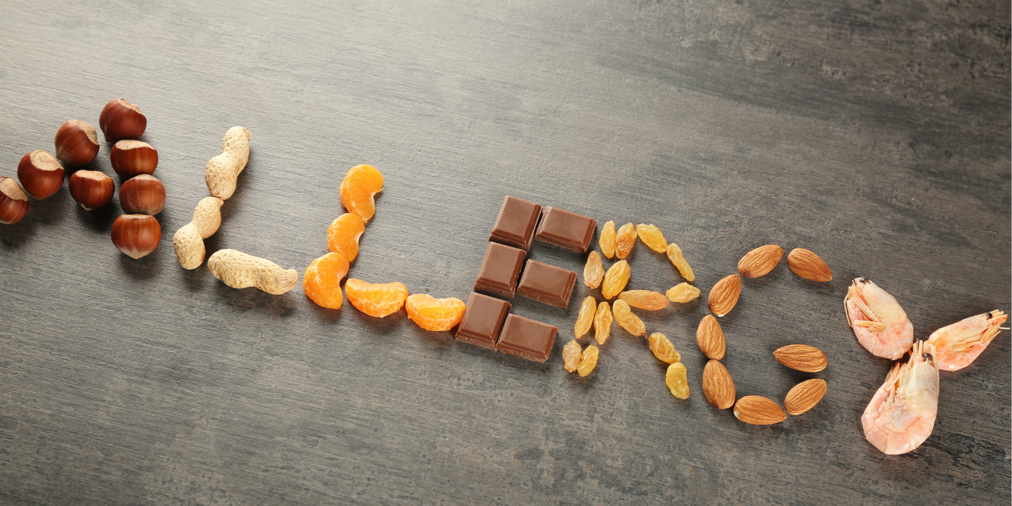 Verschiedene Lebensmittel, darunter Haselnüsse, Erdnüsse, Orangenscheiben, Schokolade, Rosinen, Mandeln und Garnelen, sind auf einer dunklen Fläche so angeordnet, dass sie das Wort "ALLERGY" ergeben.