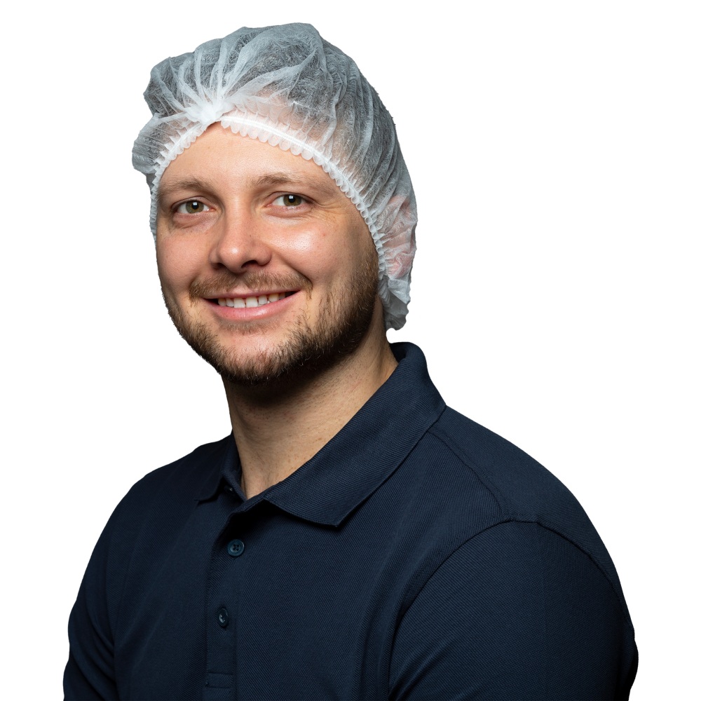 Ein Mann lächelt in die Kamera. Er trägt ein dunkelblaues Poloshirt und ein Einweg-Haarnetz NITRAS Klipphauben der MCD MEDICAL CARE DENTAL GmbH. Er hat kurze, hellbraune Haare und einen Bart vor einem schlichten weißen Hintergrund.