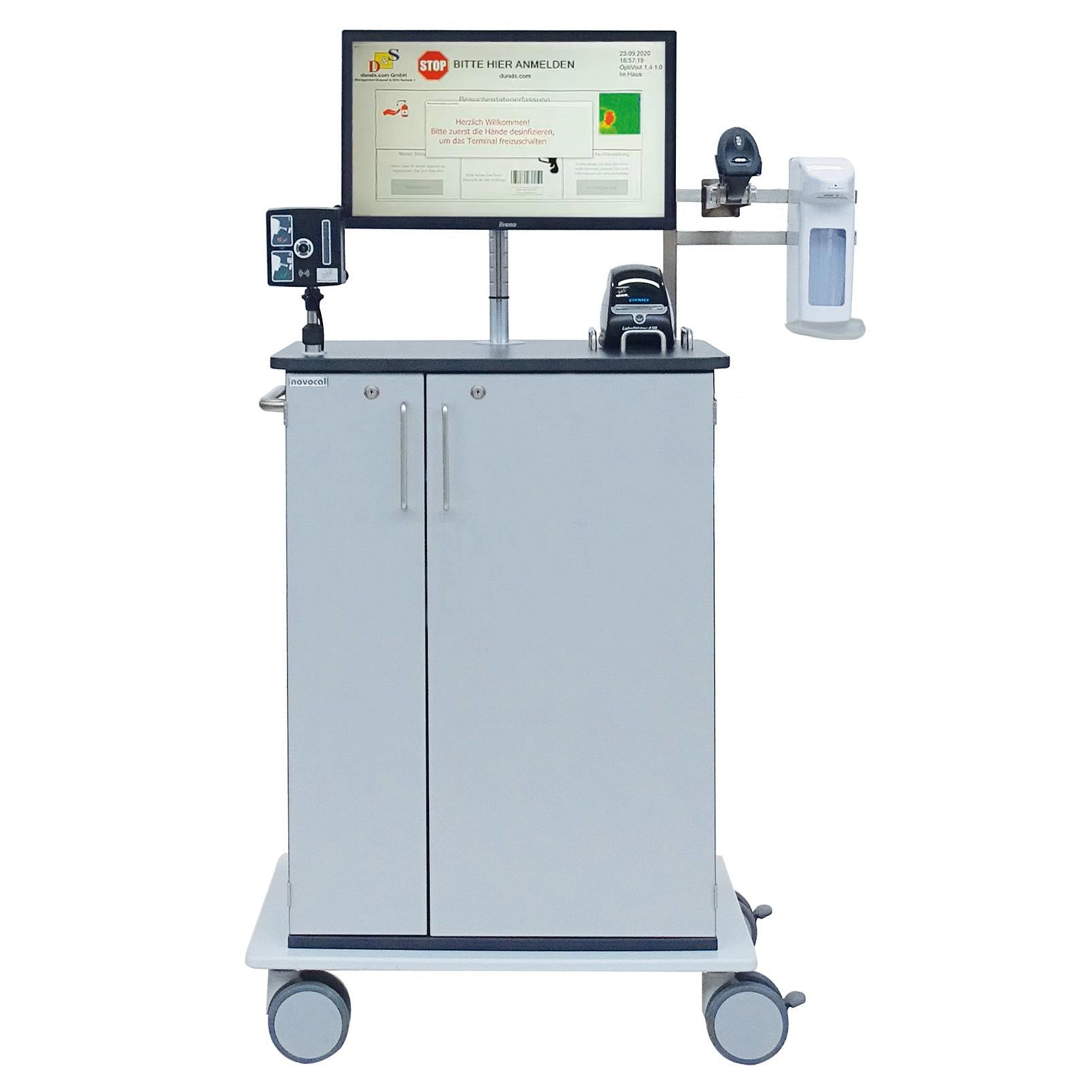 Der Novocal EDV-Wagen Typ Optivisit ist ein mobiler medizinischer Arbeitsplatz mit Monitor, Barcode-Scanner, Desinfektionsmittelspender und Drucker in einem grauen zweitürigen Schrank mit vier Lenkrollen. Marke: Novocal.
