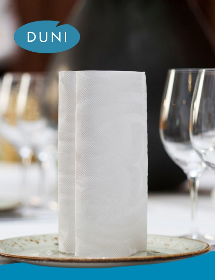 Hochformatiges DUNI-Motiv mit gefalteter weißer Serviette auf einem Teller, daneben unscharfe Weingläser im Hintergrund; oben links das DUNI-Logo, insgesamt in eleganter Tischatmosphäre.