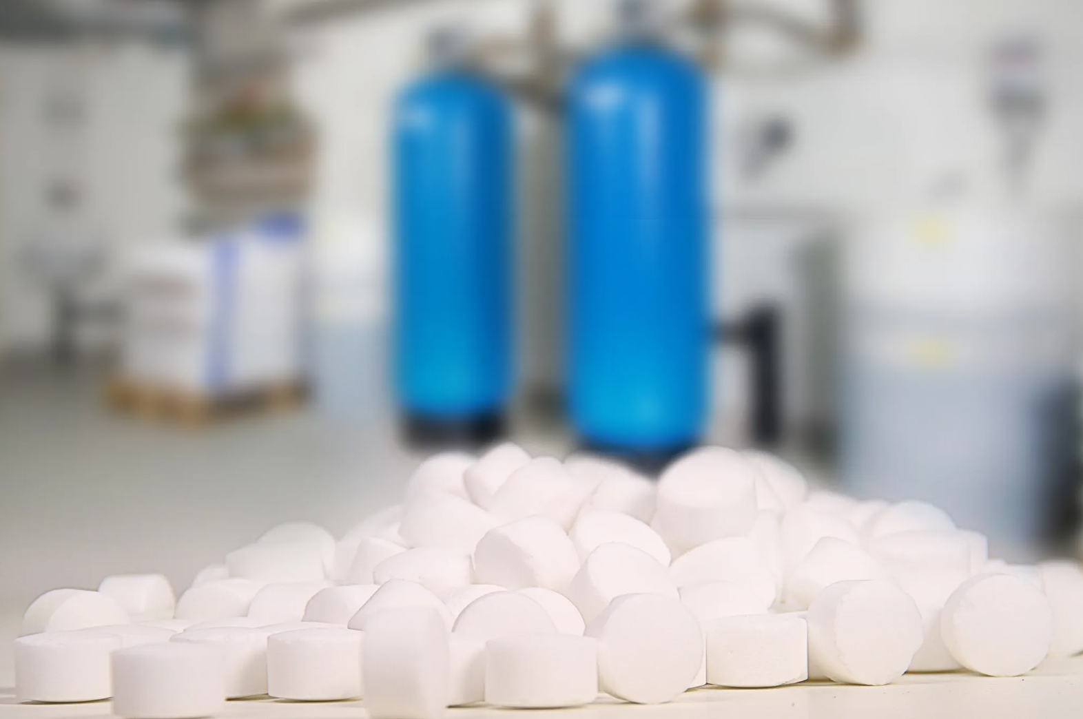 Im Vordergrund sind weiße AXAL PRO TABS scharf zu sehen, im Hintergrund unscharfe blaue Gasflaschen, in einer Industrieanlage, die auf K+S Regeneriersalz spezialisiert ist.