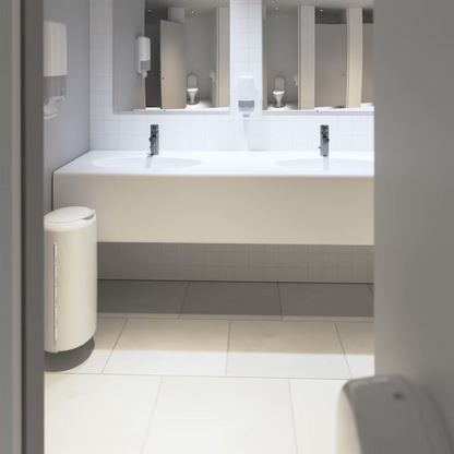 Die öffentliche Toilette präsentiert sich in modernem Elevation-Design mit weiß gefliesten Wänden und Böden. Eine lange weiße Arbeitsplatte beherbergt zwei Waschbecken unter einem großen Spiegel sowie einen Tork 553000 Spender für Zickzack-Handtücher Elevation H3 von TORK – Essity Professional Hygiene Germany GmbH, der eine effiziente Einzelblattausgabe für Papierhandtücher bietet. Ein praktisch positionierter Mülleimer befindet sich in der Nähe des Eingangs.