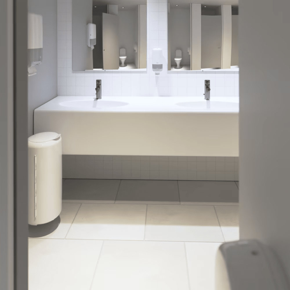 Man sieht eine saubere, moderne öffentliche Toilette mit einer langen weißen Theke mit zwei Waschbecken und Wasserhähnen. Unter der Theke fällt eine auffällige weiße Fliesenwand auf. Der Raum ist mit Spiegeln, Tork-Seifenspendern von Essity Professional Hygiene Germany GmbH und einem weißen Mülleimer auf einem Fliesenboden ausgestattet, der für stark frequentierte Toiletten konzipiert ist.