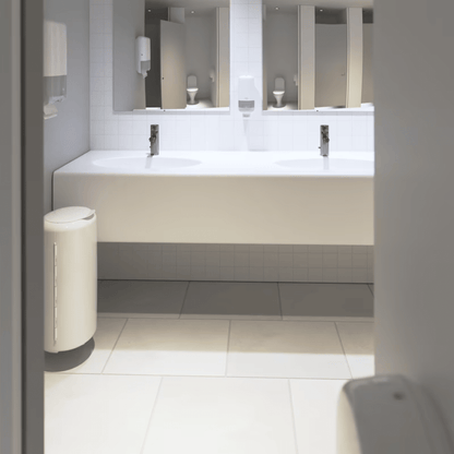 Man sieht eine saubere, moderne öffentliche Toilette mit einer langen weißen Theke mit zwei Waschbecken und Wasserhähnen. Unter der Theke fällt eine auffällige weiße Fliesenwand auf. Der Raum ist mit Spiegeln, Tork-Seifenspendern von Essity Professional Hygiene Germany GmbH und einem weißen Mülleimer auf einem Fliesenboden ausgestattet, der für stark frequentierte Toiletten konzipiert ist.